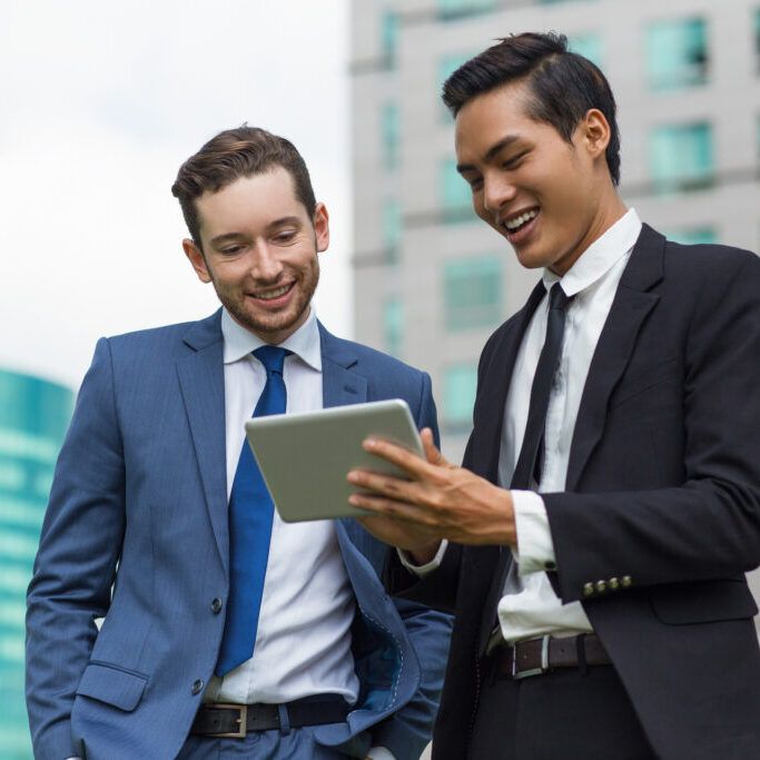 Closeup of two smiling young business men using tablet computer and standing with office buildings in background