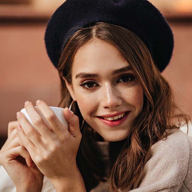 pretty-young-lady-with-brunette-wavy-hair-dark-eyes-trendy-makeup-beret-pullover-sitting-outdoors-with-cup-tea-smiling